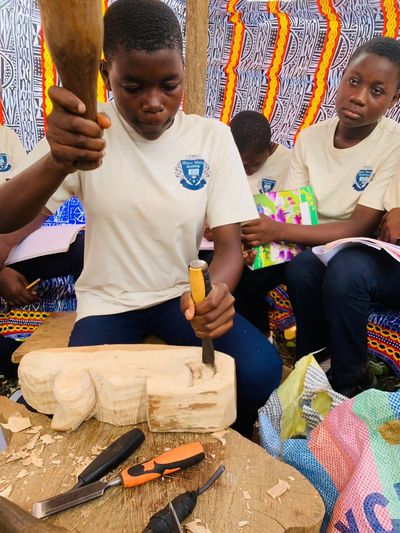 A student carving a wooden form with a mallet and chisel while classmates take notes