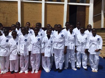 Students in white martial arts uniforms with the school crest, standing on training mats