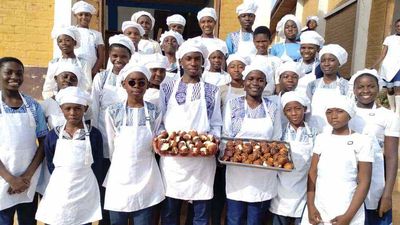 A large group of students in chef whites holding trays of baked goods