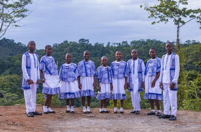 Eight Mfàh’Mfàh Academy students in blue-and-white uniform standing together on a ridge above rainforest