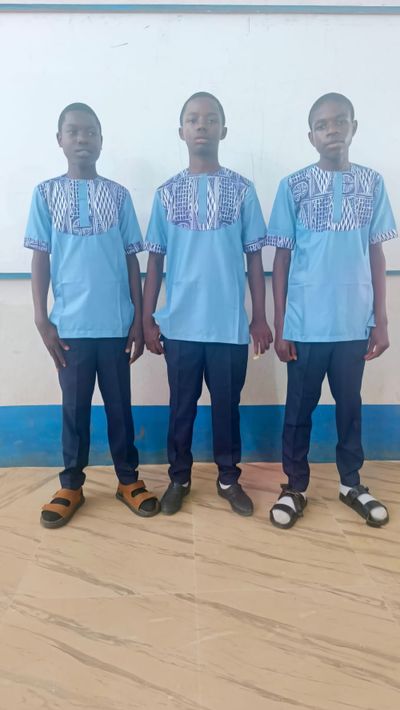 Three students in the day uniform for boys: a light blue tunic with an Ndop-patterned yoke and cuffs, worn with navy trousers