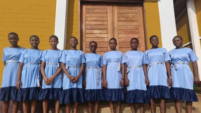 Nine students in the light blue day uniform with a navy hem and an Ndop-patterned sash, standing together outside a school building